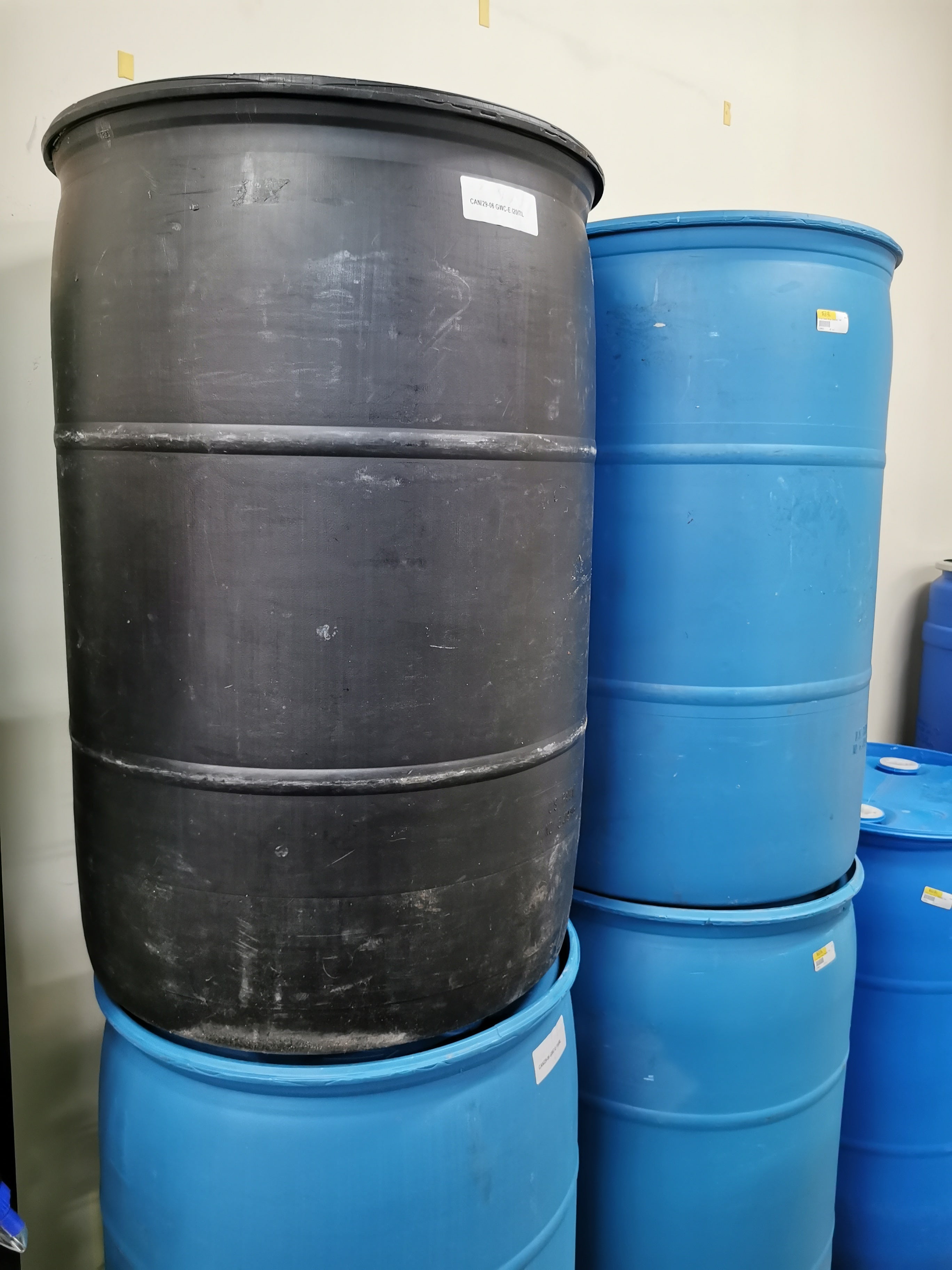 A black reconditioned 55 gallon plastic drum with visible signs of wear, stacked among other drums.