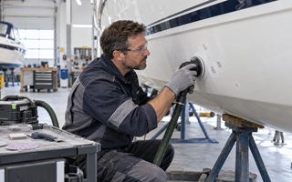 Marine repair technician sanding localized gelcoat blisters on a white fiberglass boat hull with dust extraction