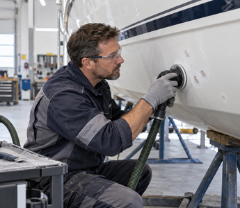 Marine repair technician sanding localized gelcoat blisters on a white fiberglass boat hull with dust extraction